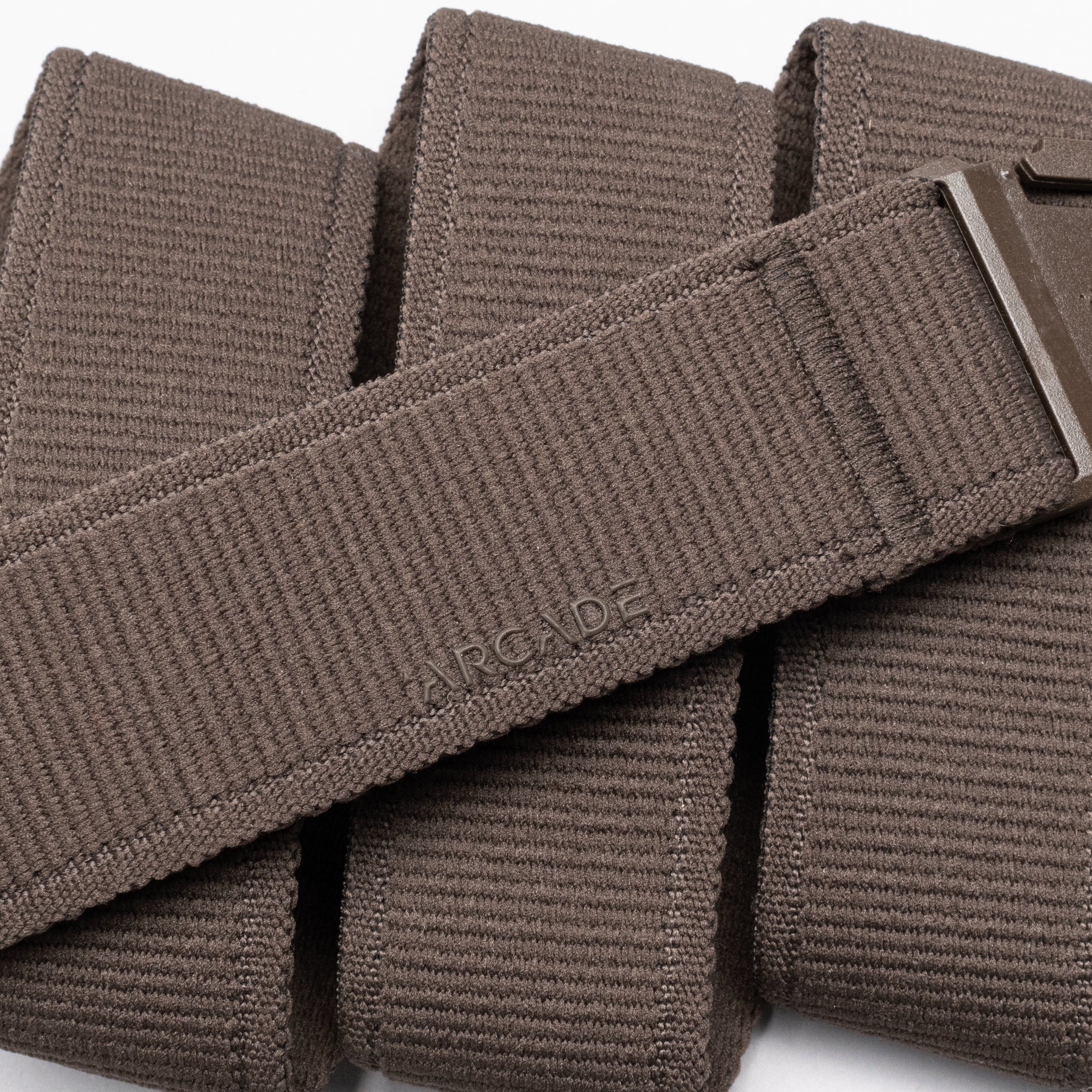 A close-up of the Atlas belt by Arcade Belts, featuring brown woven stretch webbing, a low-profile metal buckle, and a subtly embossed ARCADE logo on the folded strap—highlighting the brand's signature style.