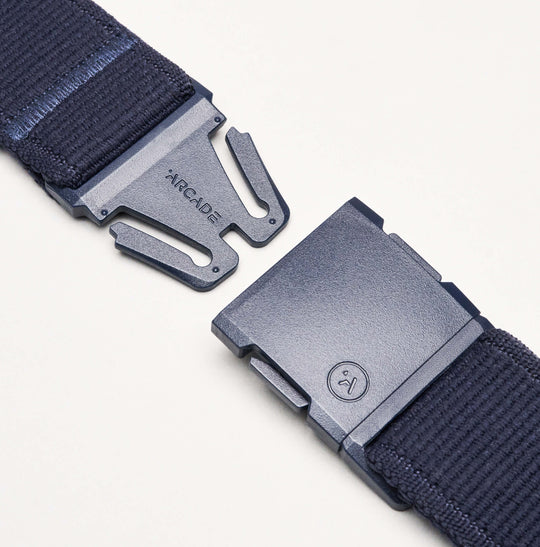 A close-up of the navy blue Atlas Elastic Stretch Belt by Arcade Belts, featuring a metal clasp that's partially unbuckled, with ARCADE etched on one side and a small circular logo on the other, showcasing its quality craftsmanship.
