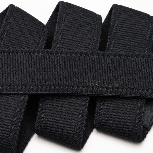 A close-up shows the Atlas belt by Arcade Belts, black with ARCADE subtly embossed on its woven surface. The neatly folded belt sits on a white background, highlighting its comfortable stretch webbing.