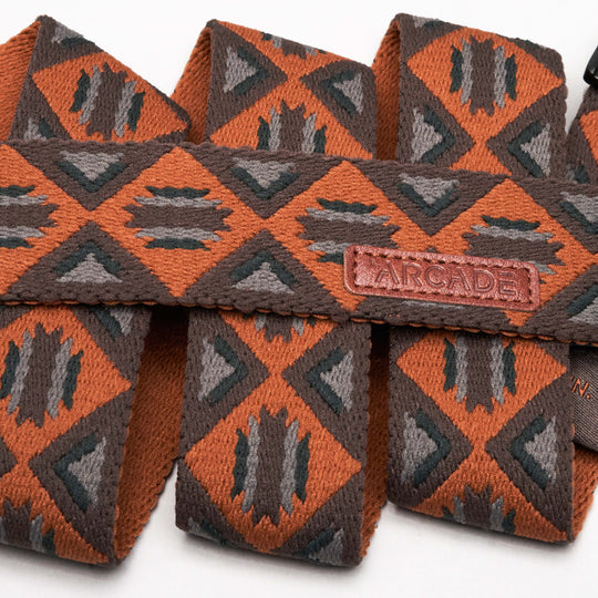 A close-up of the Vernan Kee belt by Arcade Belts, showcasing orange and brown geometric patterns, a brown leather Arcade tag, and a sturdy plastic buckle—perfect as a stylish travel accessory.