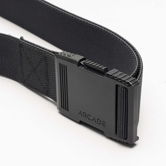A close-up of the Arcade Belts Hardware: a black elastic stretch belt with a rectangular black plastic buckle engraved with "ARCADE," shown against a white background.