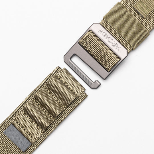 Close-up of a tan nylon Arcade Belts Hardware Alu belt with a rectangular metal buckle engraved "ARCADE." The stretch webbing strap features stitched loops and a reflective strip, displayed on a white background.
