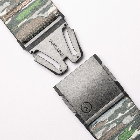 Close-up of the Realtree Camo Original by Arcade Belts, a stretch webbing belt with a camouflage pattern, partially open metal buckle engraved "ARCADE," and a small lower logo.