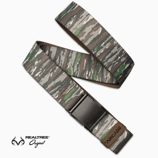 The Arcade Belts Realtree Camo Original features a black buckle, Arcade logo on one end, and Realtree Original logo in the bottom left. Its camo pattern mixes green, brown, gray, and beige for an authentic outdoor style.